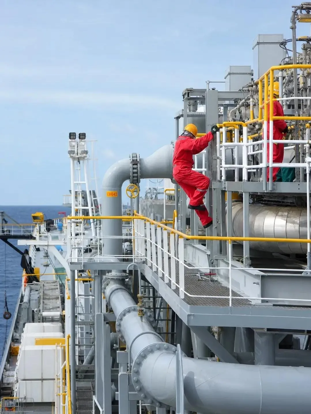Offshore crew in red coveralls climbing pipework on an LNG carrier deck at sea